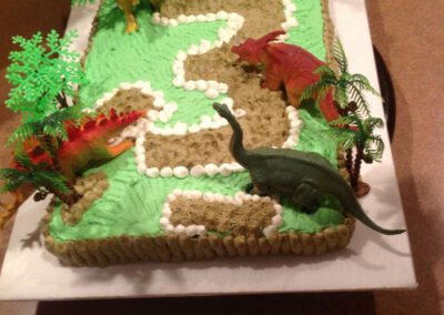 A themed cake decoration featuring a dinosaur and miniature trees. The cake is designed with a dinosaur motif, complete with small trees to create a prehistoric scene