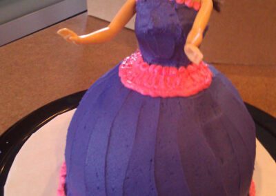 A cake designed to look like a girl, featuring a dress in pink and purple colors with cream accents. The cake is decorated to resemble a girl's outfit, making it a charming and whimsical dessert